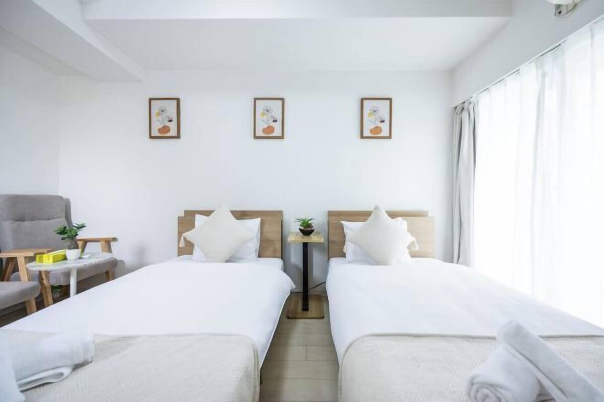 Une chambre d’hôtel avec deux lits simples, une petite table avec une plante, une chaise et une fenêtre avec des rideaux.