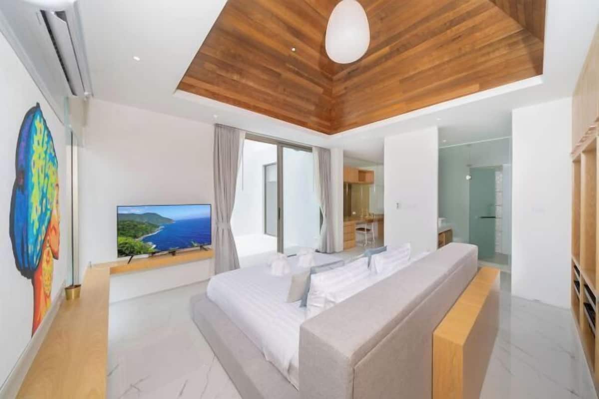 A modern bedroom with a wooden ceiling, a large bed, a TV on the wall, and a view of the sea.