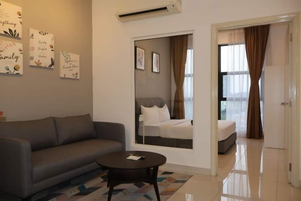 Une chambre d’hôtel moderne avec un canapé gris, une petite table ronde et un lit avec une tête de lit. Un grand miroir orne le mur, et la chambre présente un design épuré et minimaliste.