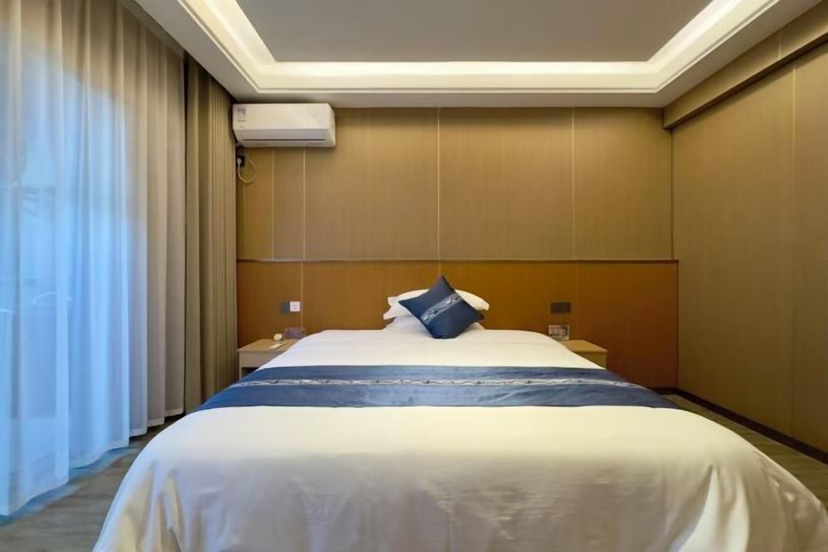 A hotel room with a bed, curtains, an air conditioning unit, and a nightstand.