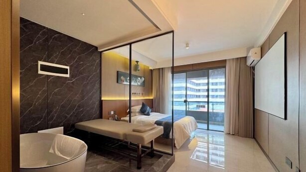 A modern hotel room with a large bathtub, a bench, a bed, a TV, and a balcony with a view of the city. - Boten Jinchi holiday Spa Hotel (Boten)