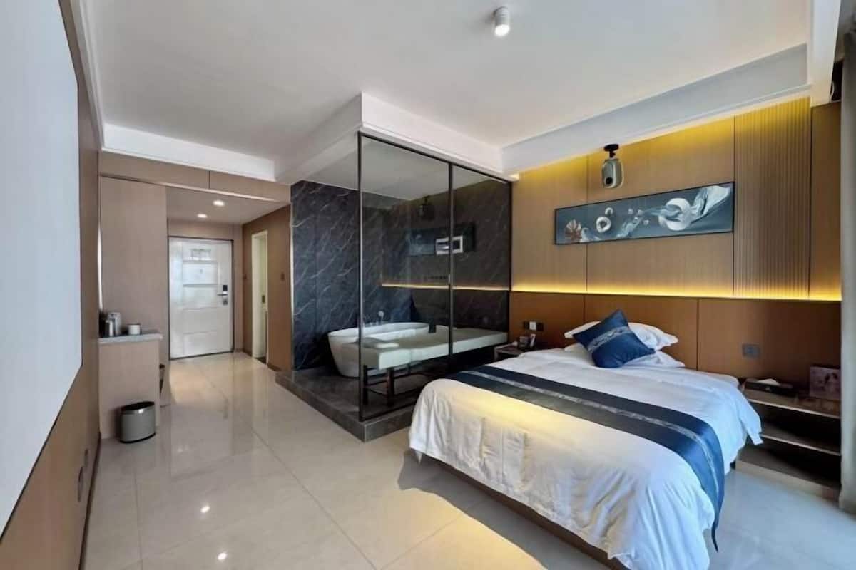 A modern hotel room with a large bed, a glass-enclosed bathroom, and a wall-mounted artwork.