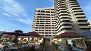 A tall hotel with multiple balconies, outdoor seating areas, and a central pathway leading to the entrance. - Boten Jinchi holiday Spa Hotel (Boten)
