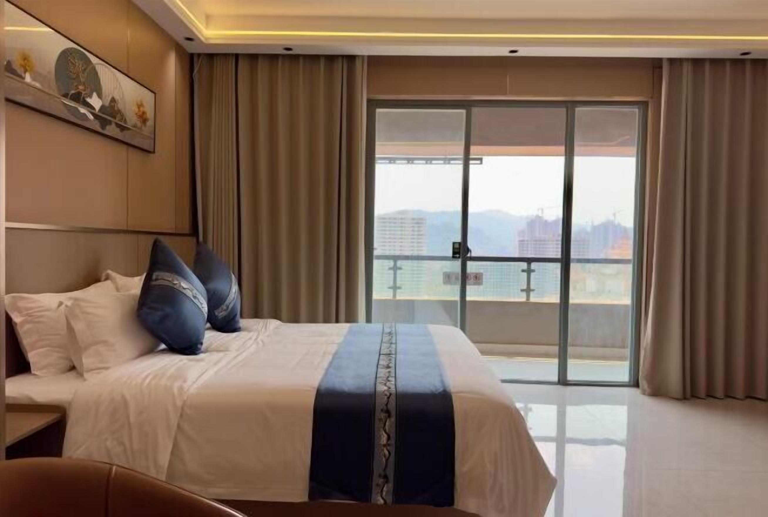 A hotel room with a bed, a view of buildings through a large window, and a door to the outside.