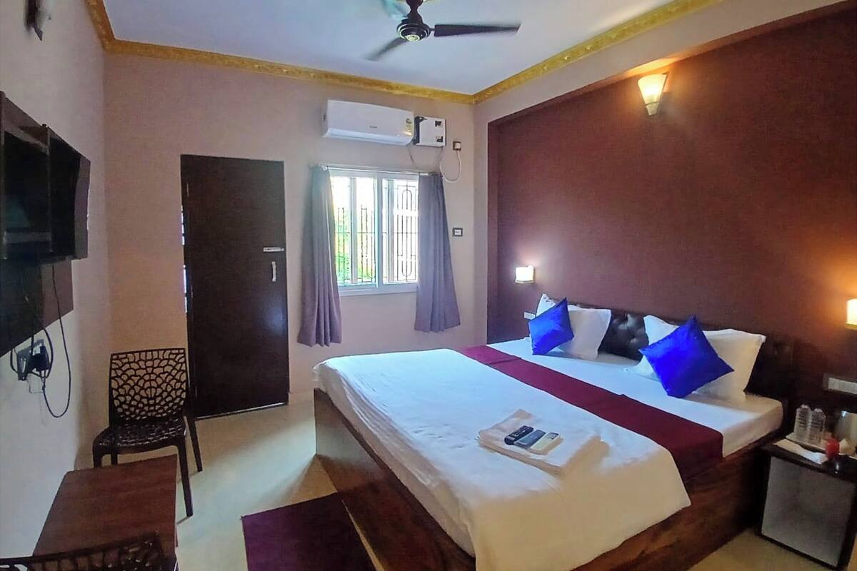 Habitación de hotel con cama, escritorio, silla, televisor, aire acondicionado, ventilador de techo y ventana con cortinas.