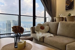 Ein modernes Wohnzimmer mit einem großen Fenster, einem beigefarbenen Sofa, einer Vase mit getrockneten Blumen und einem Holztisch mit Kerzen.