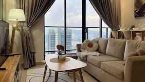 A modern living room with a sofa, a small table with a vase, and a TV on a wooden stand. Large windows offer a city view.