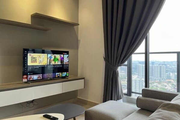 Un salon moderne avec un téléviseur à écran plat fixé au mur, un canapé, une petite table et une vue sur le paysage urbain à travers de grandes fenêtres.