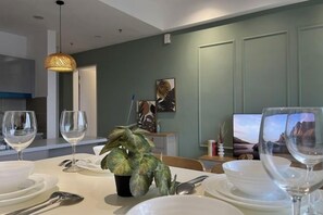 Un área de comedor con una mesa lista para dos personas, una planta en maceta y un televisor al fondo.