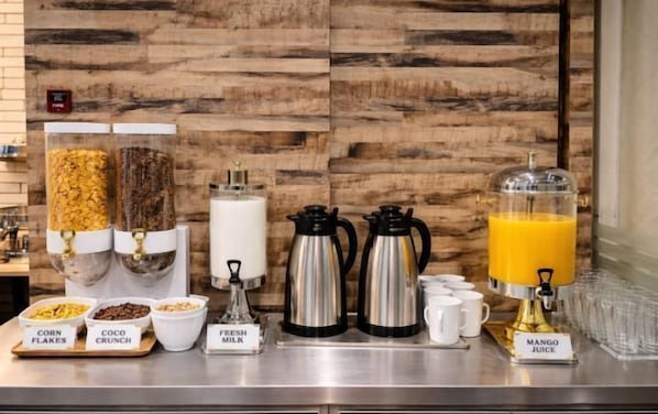 Una barra de desayuno con recipientes etiquetados de copos de maíz, coco crujiente y leche fresca, junto con cafeteras y un dispensador de jugo.
