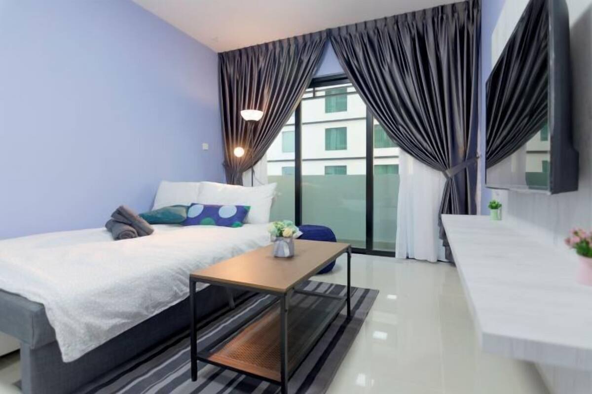 A modern hotel room with a bed, a small table, a desk, and a view of the city through large windows.