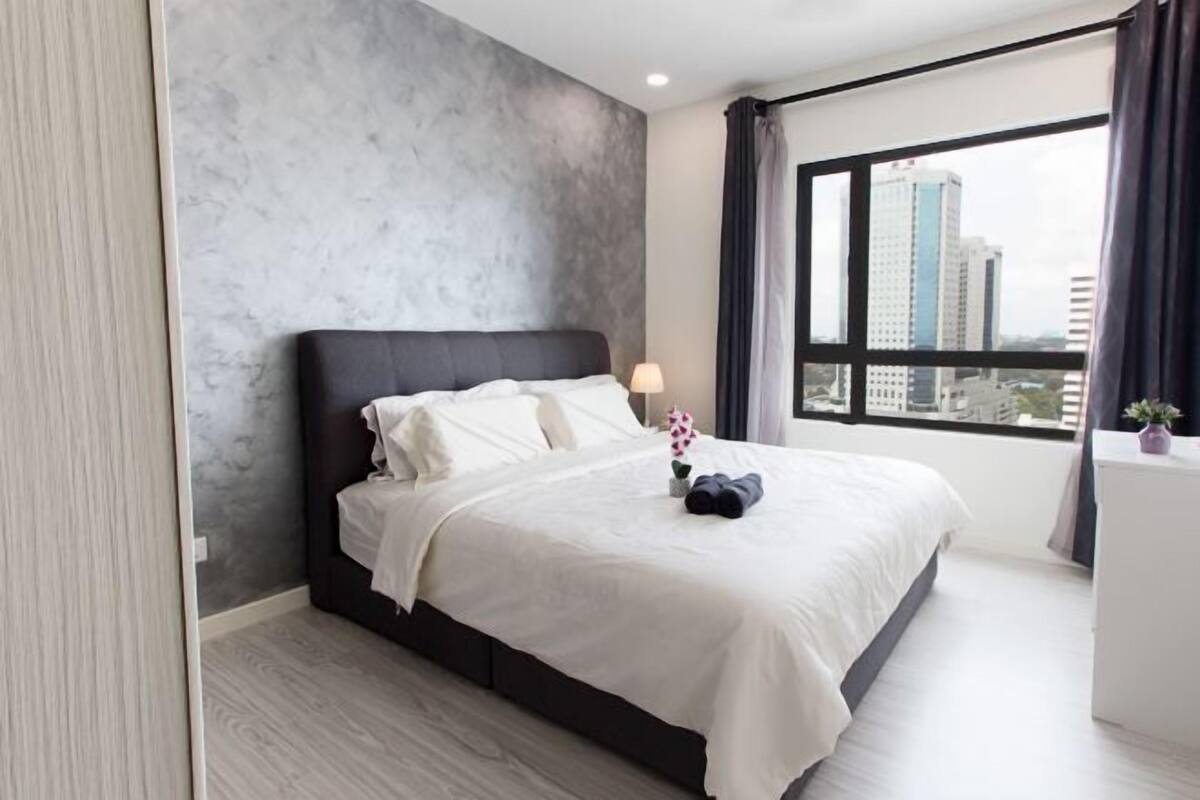 A modern hotel room with a large bed, a city view, and a grey textured wall.