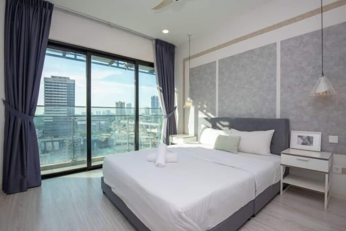 A modern hotel room with a large bed, a bedside table, a nightstand, and a view of the city through floor-to-ceiling windows.