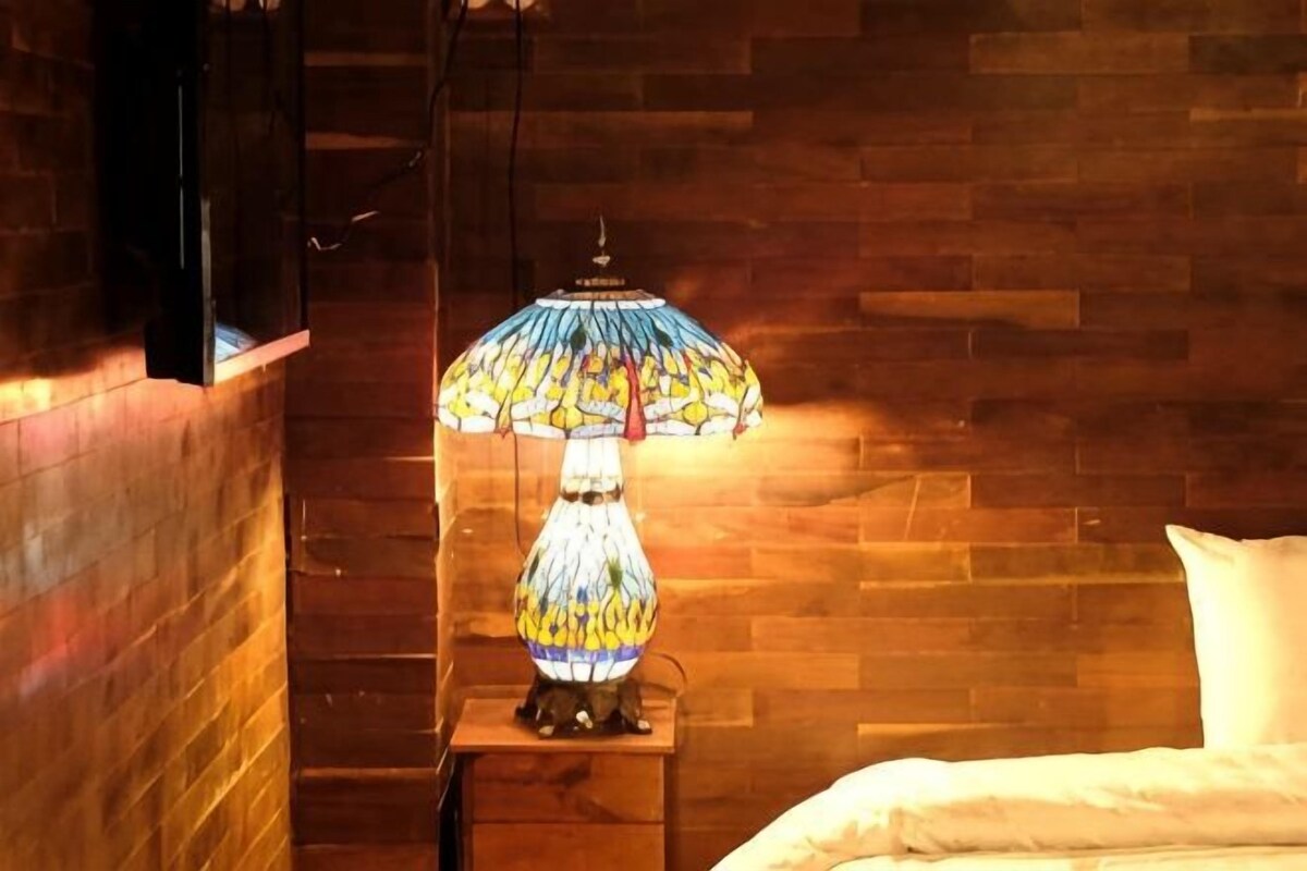 A room with a wooden wall, a bedside table with a lamp, and a bed with pillows.