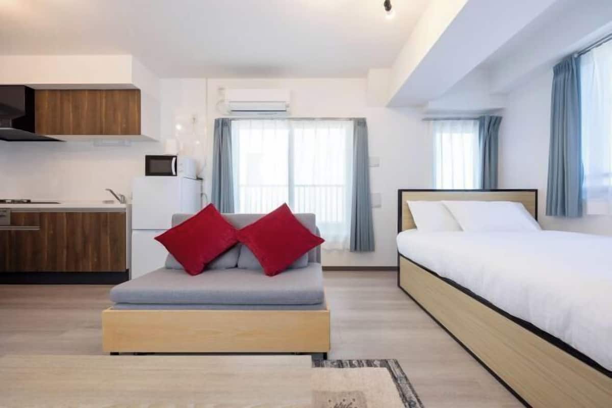 A modern hotel room with a kitchenette, a sofa with red cushions, a bed, and large windows with curtains.
