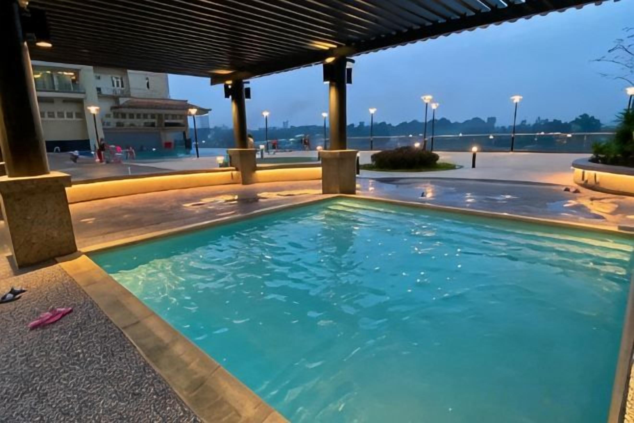 Una piscina amb una zona coberta, envoltada per un passeig pavimentat i un edifici al fons.