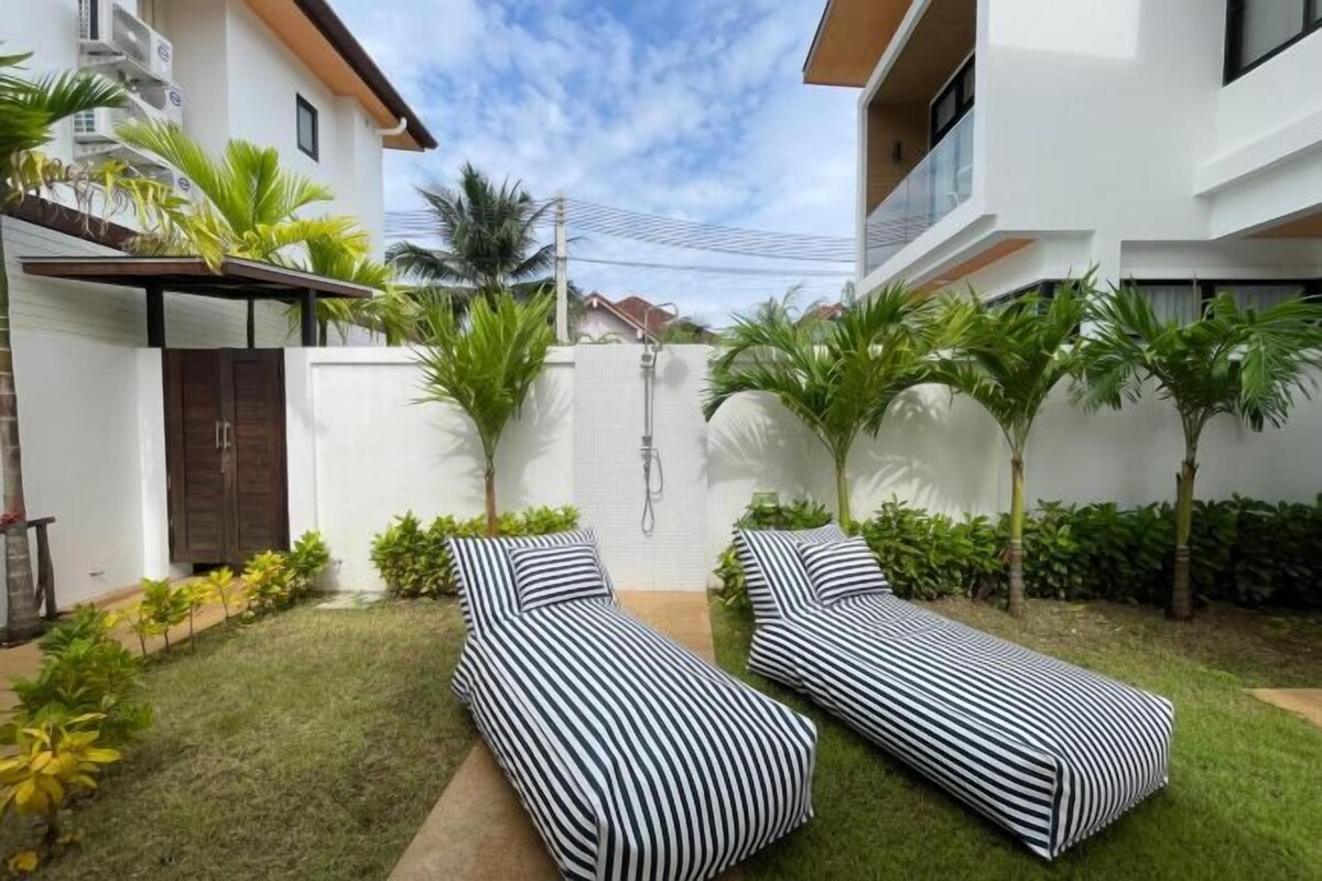 A modern outdoor seating area with two lounge chairs, a white wall, and a small garden with palm trees.
