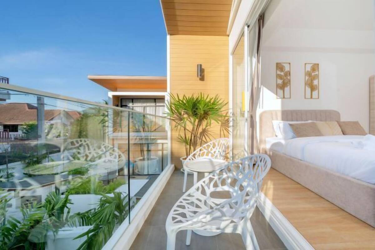 Een balkon met glazen hekwerk, witte stoelen, een klein tafeltje en een bed met een beige hoofdeinde.