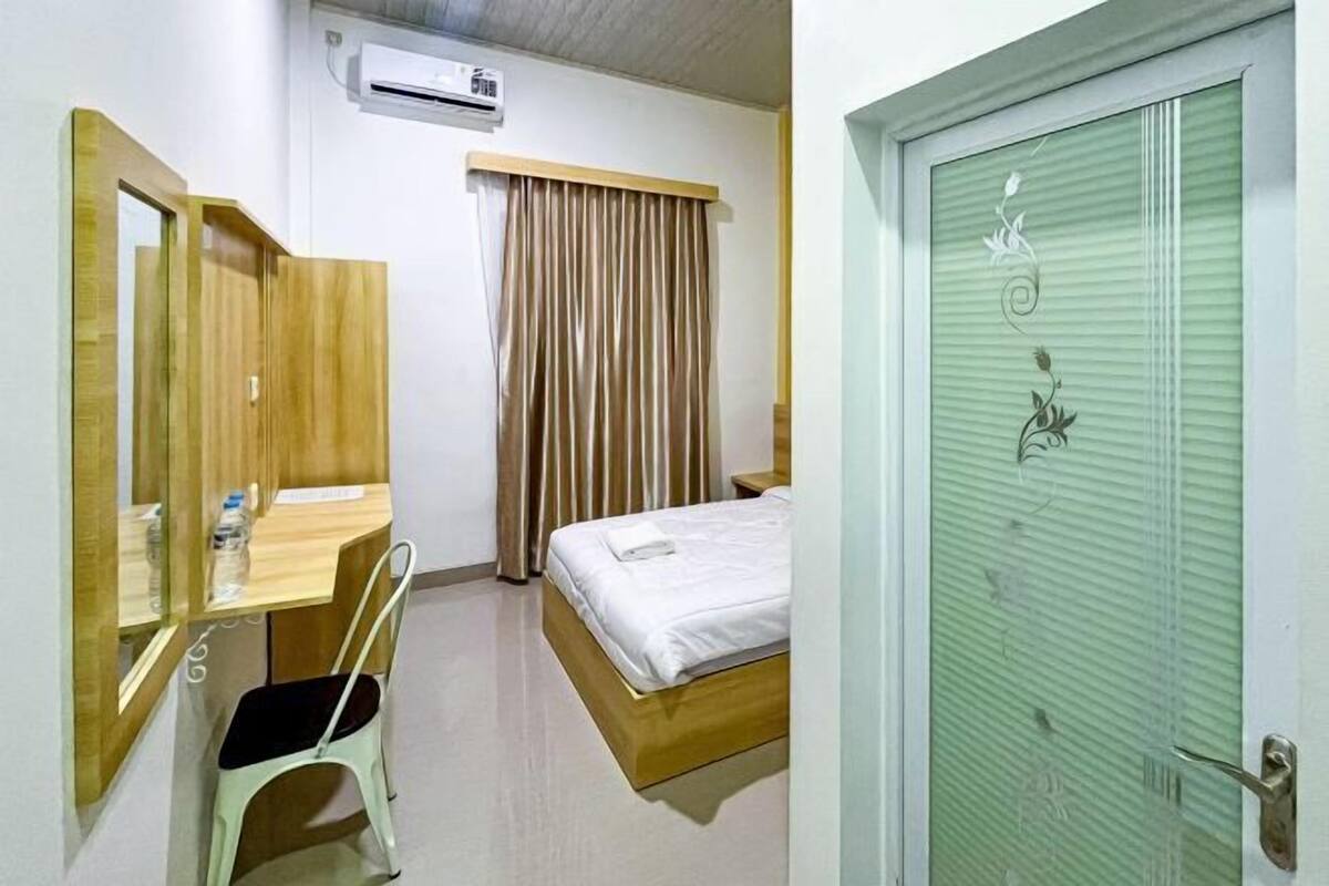 A hotel room with a wooden desk, a chair, a bed with a white mattress, a window with curtains, and a green door with a floral design.