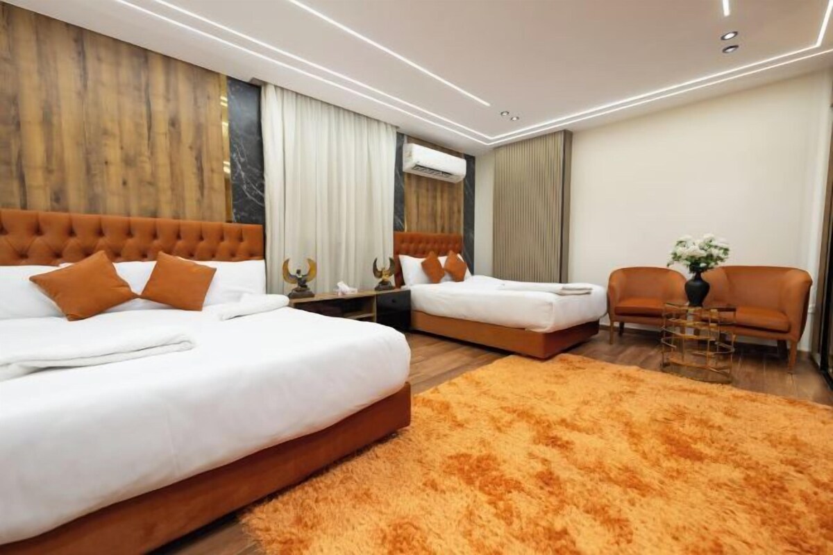 A modern hotel room with two beds, a sofa, a coffee table, and a large orange rug.