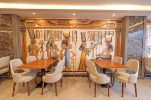 Un comedor con un mural de figuras egipcias, mesas de madera y sillas blancas.