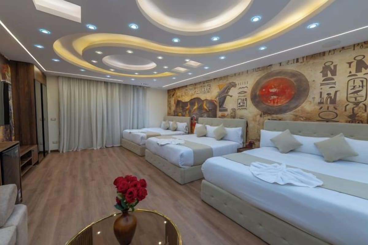 A hotel room with three beds, a wooden floor, a wall mural, and a vase with red flowers.