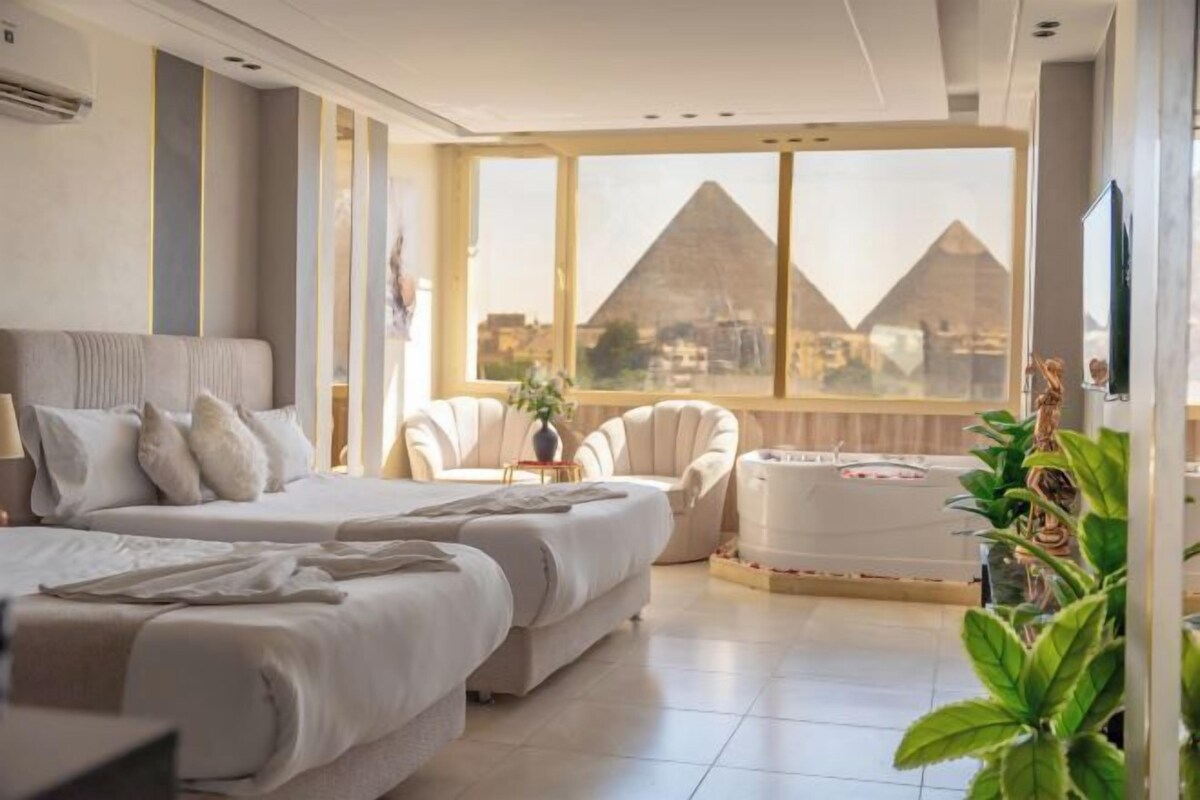 A hotel room with a bed, two chairs, a small table, and a view of the pyramids.