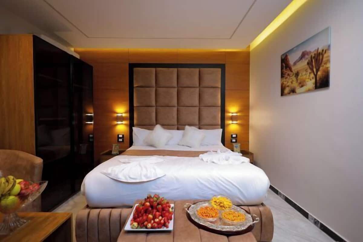 Una habitació d'hotel amb un llit gran, làmpades de nit, un panell de fusta al capçal, una taula de menjador amb fruita i un quadre amb temàtica de desert.