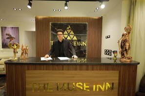 A reception desk with a logo, a man seated behind it, and a statue on the right. - The Muse Pyramids INN (Kafr Nassar)