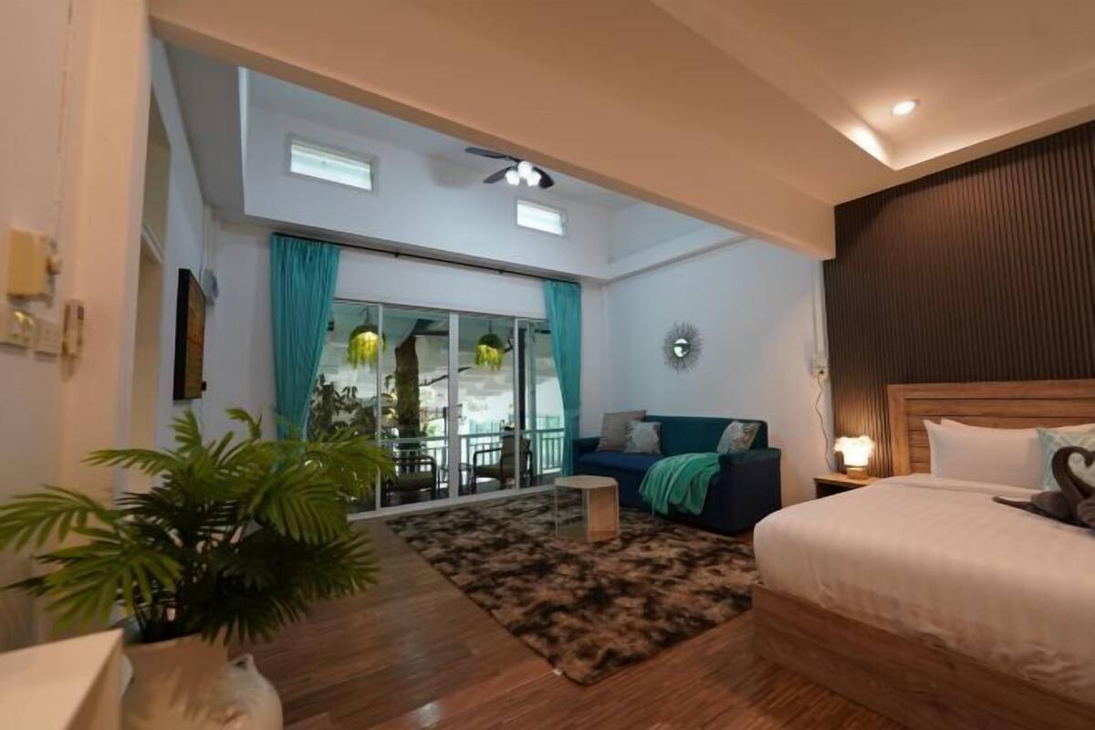 A hotel room with a bed, a sofa, a small table, a potted plant, a view of the outside, and a ceiling fan.