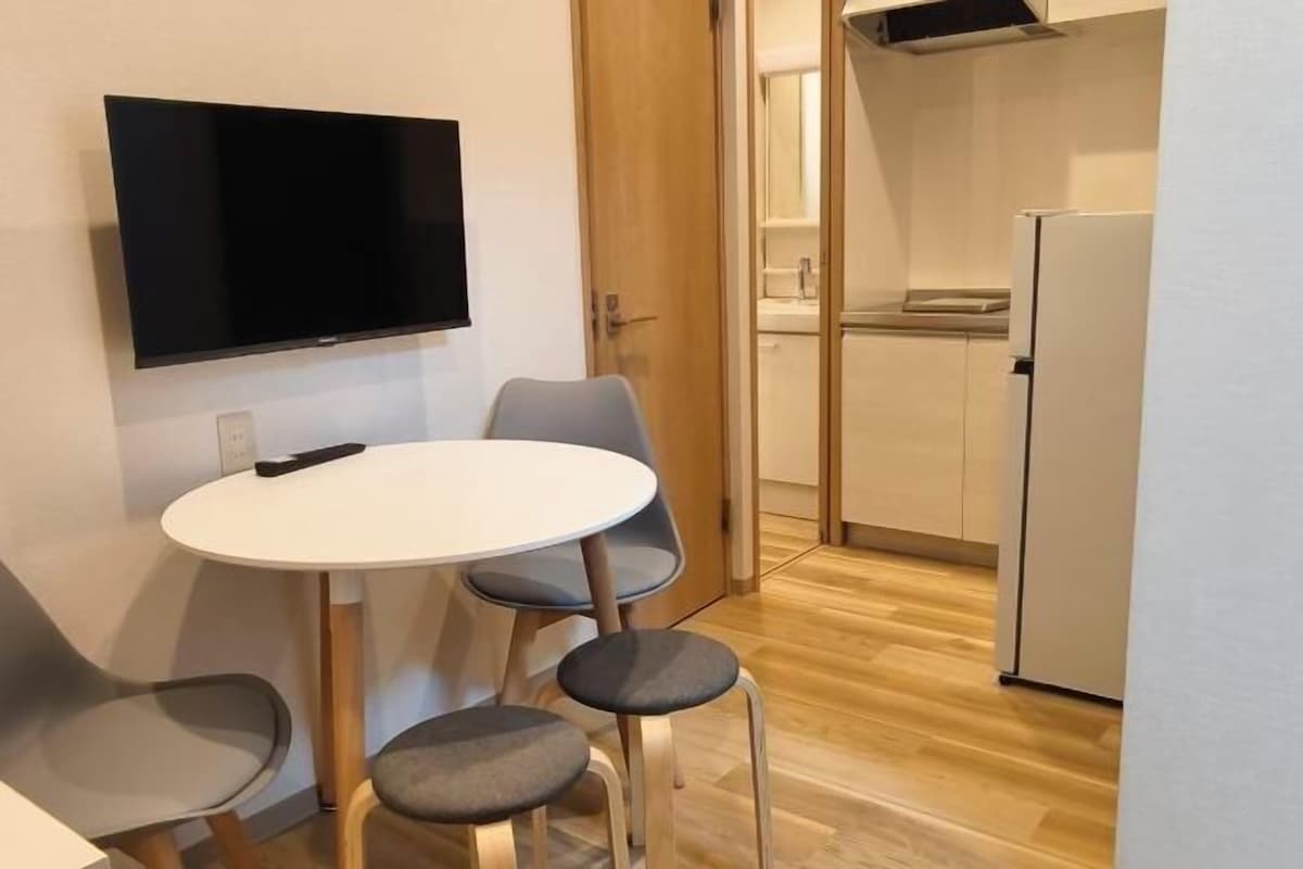 Una habitación pequeña con televisión, una mesa redonda, dos sillas, una puerta que da a la cocina y un refrigerador.