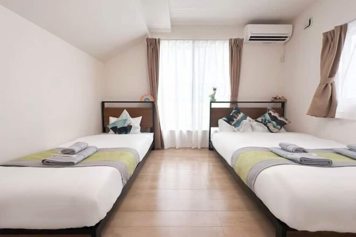 Habitación de hotel con dos camas individuales, una ventana con cortinas, un equipo de aire acondicionado y una pequeña mesa con una planta.