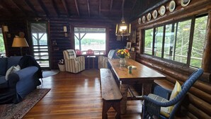 Interior rústico de una cabaña con una mesa de comedor de madera, sillas tapizadas en azul, un banco y un ventanal con vistas.