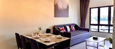 A dining area with a table set for four, a sofa with cushions, a coffee table, and a large window with curtains.