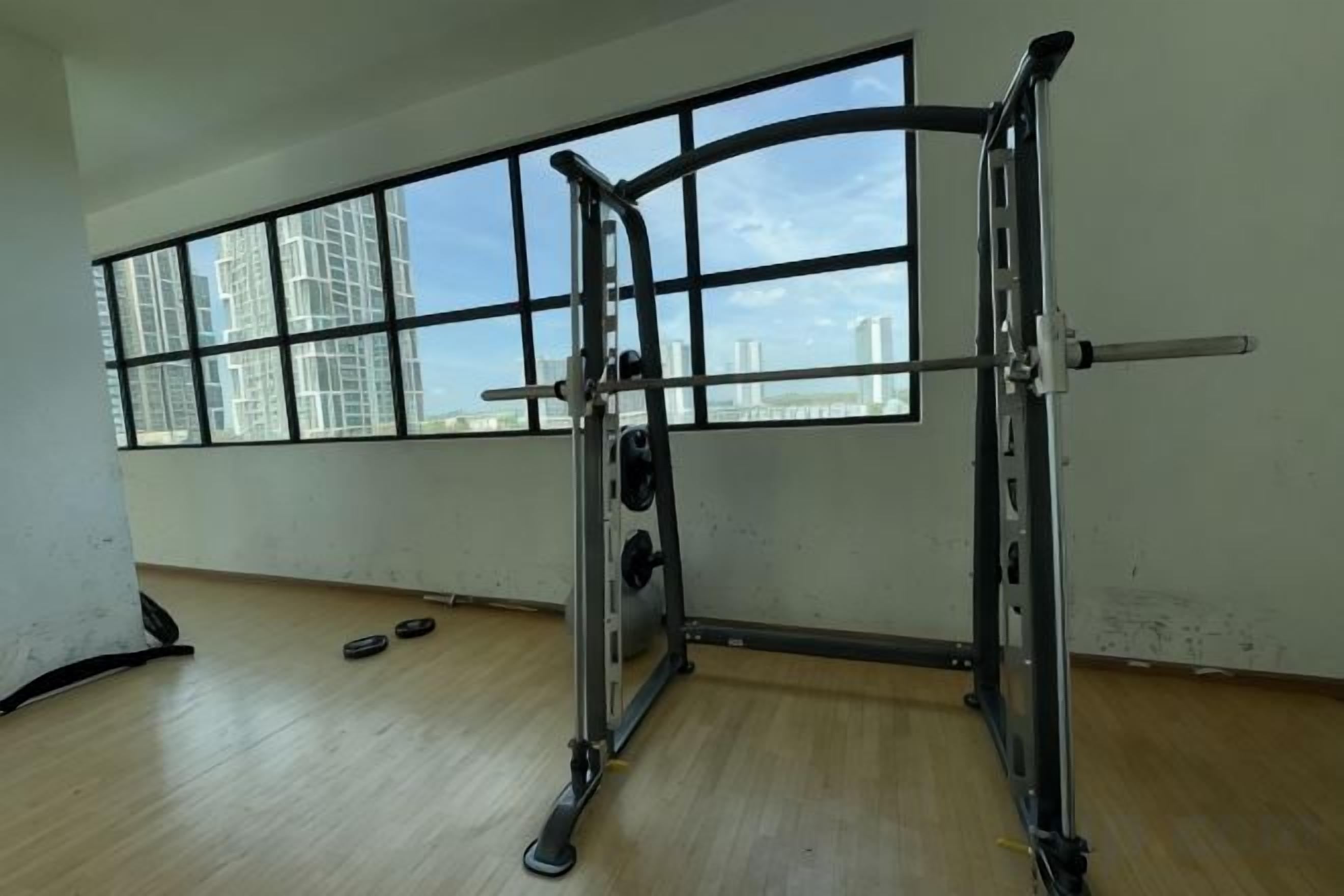 Een fitnessruimte met een power rack, een bankdrukpers en een stang. Er zijn ramen met uitzicht op gebouwen.