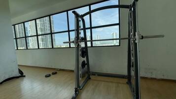 Een fitnessruimte met een power rack, een bankdrukpers en een stang. Er zijn ramen met uitzicht op gebouwen.