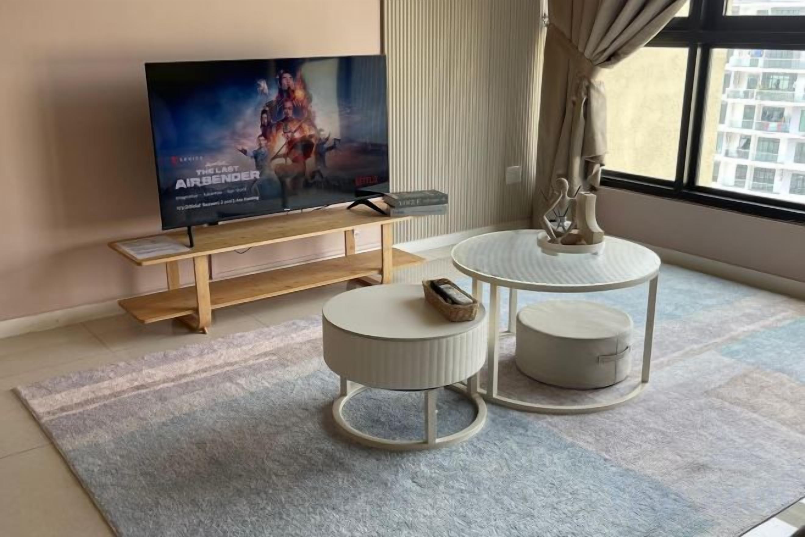 A modern living room with a flat-screen TV, a wooden coffee table, two round side tables, and a large window.