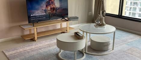 A modern living room with a flat-screen TV, a wooden coffee table, two round side tables, and a large window.