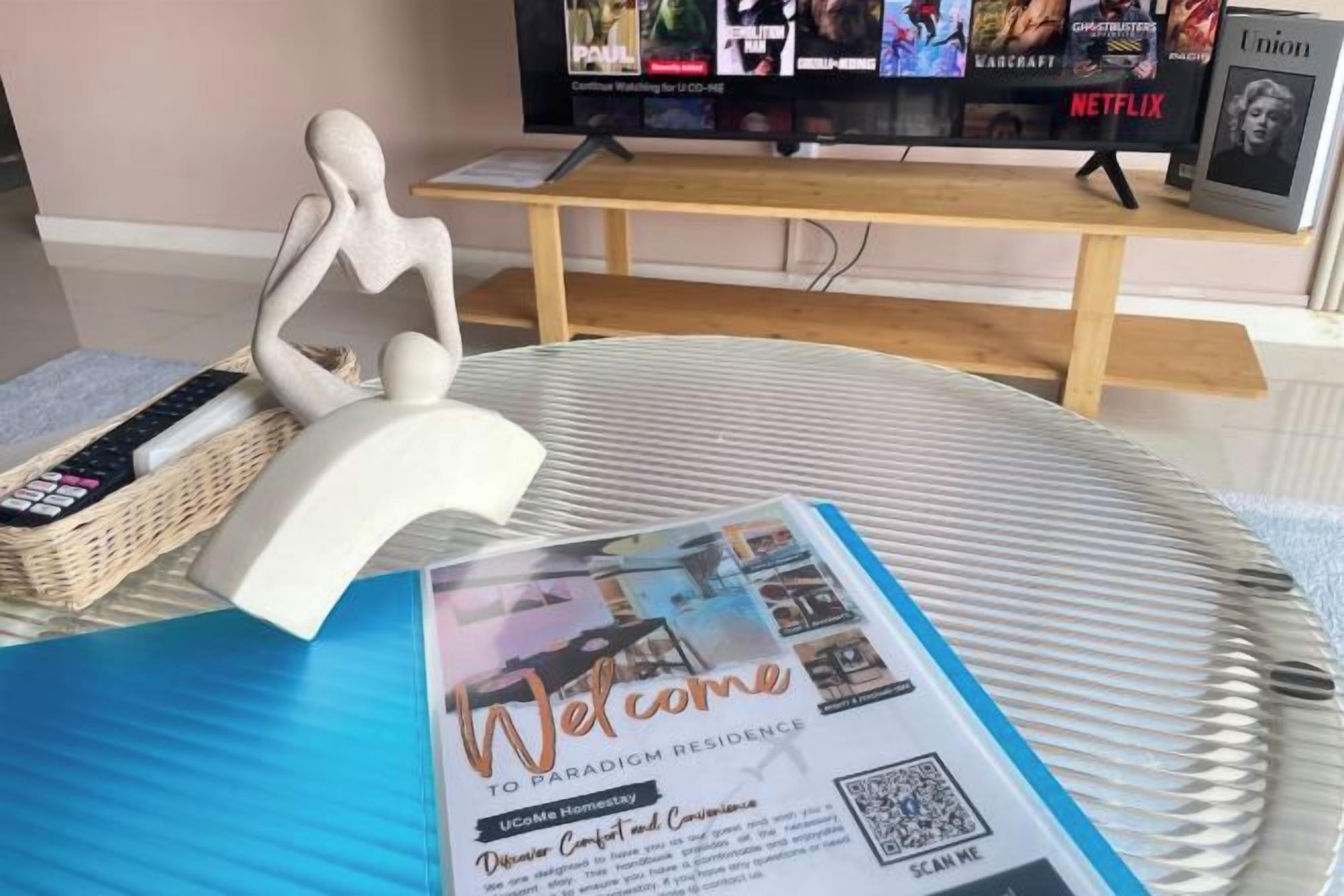 Une brochure d’accueil est posée sur une table, à côté d’une télécommande et d’un objet décoratif. Un téléviseur diffusant différentes chaînes est visible à l’arrière-plan.