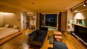 A modern hotel room with a sofa, a TV, a dining table, and a large window with a city view.