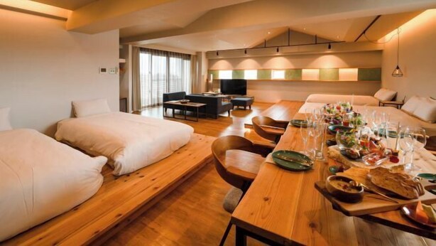 A hotel room with a wooden dining table set with food and drinks, a sofa, a TV, and a large window.