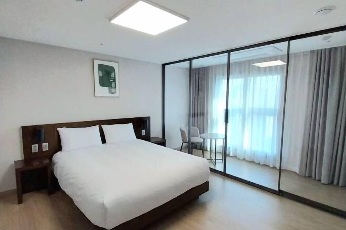 A hotel room with a bed, a bedside table, a chair, a small table, and a sliding glass door leading to a balcony.
