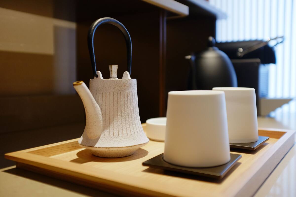 A white ceramic kettle with a blue handle on a wooden tray, accompanied by two white ceramic cups.