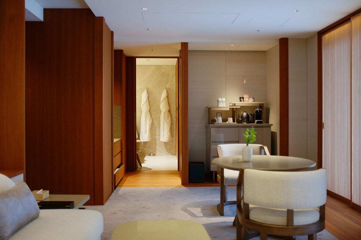A hotel room with a wooden wardrobe, a seating area with a table and chairs, a small kitchenette, and a bathroom visible through an open door.