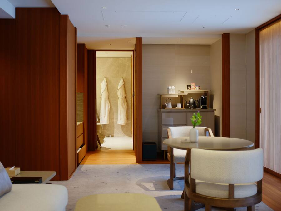 A hotel room with a wooden wardrobe, a seating area with a table and chairs, a small kitchenette, and a bathroom visible through an open door.
