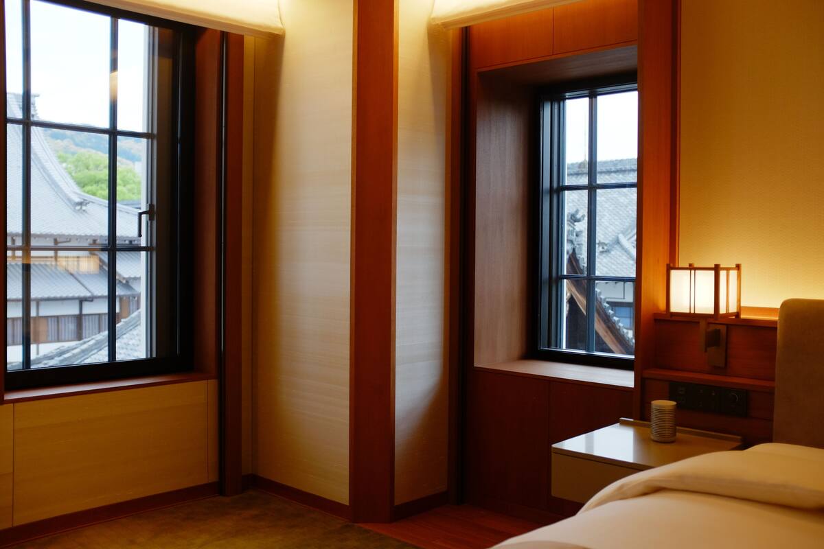 A hotel room with a large window, a bedside table with a lamp, and a bed.