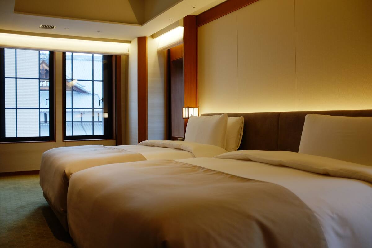 A hotel room with two beds, a large window, and a wall-mounted lamp.