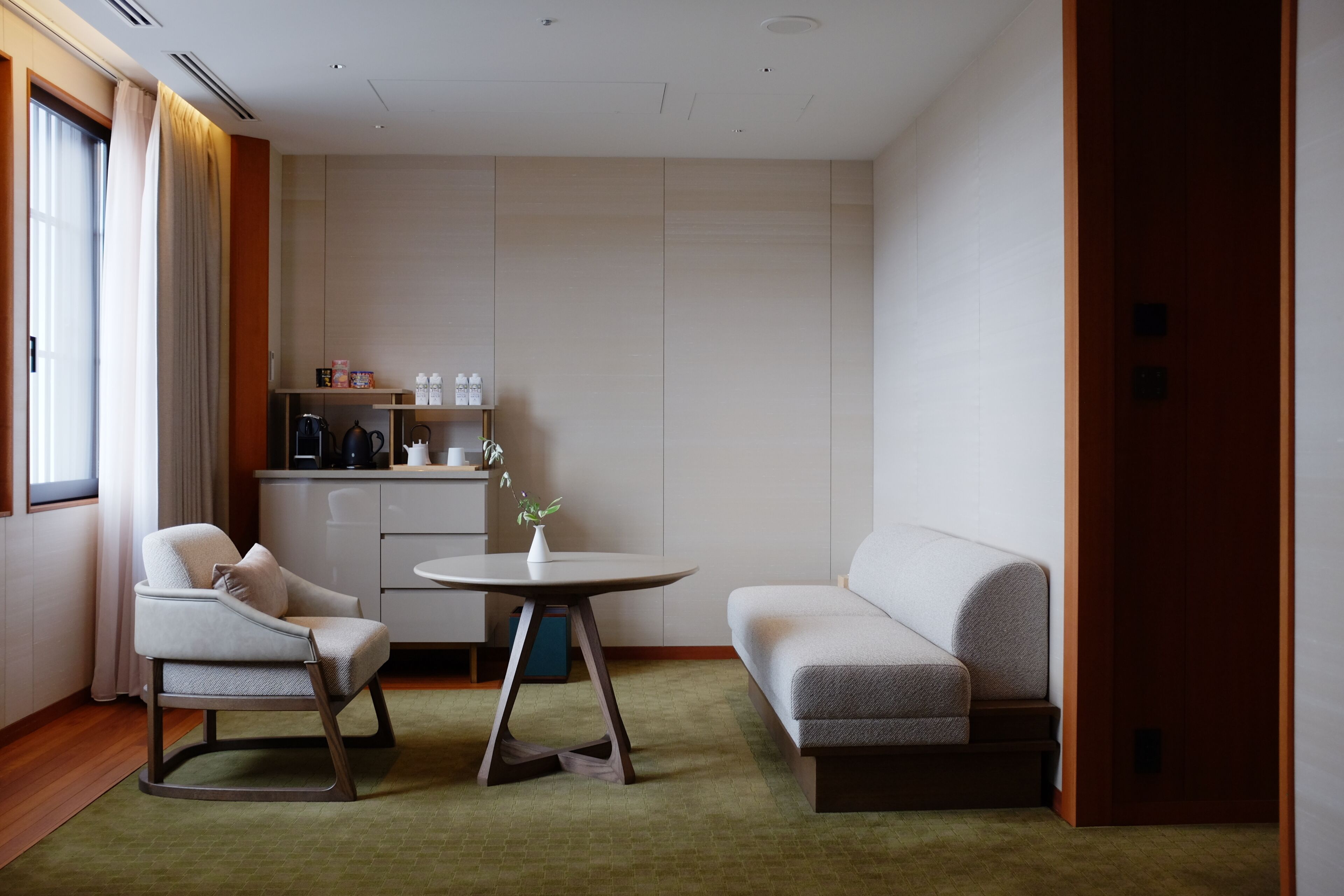 Una habitación de hotel moderna con sofá, una silla, una mesita, kitchenette y una ventana con cortinas.