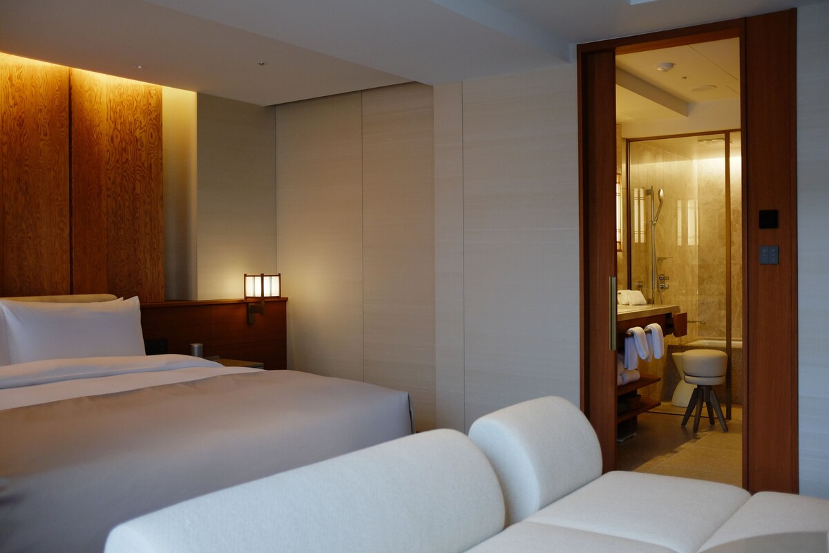Habitación de hotel con cama, sofá, pared de paneles de madera y baño visible a través de una puerta abierta.