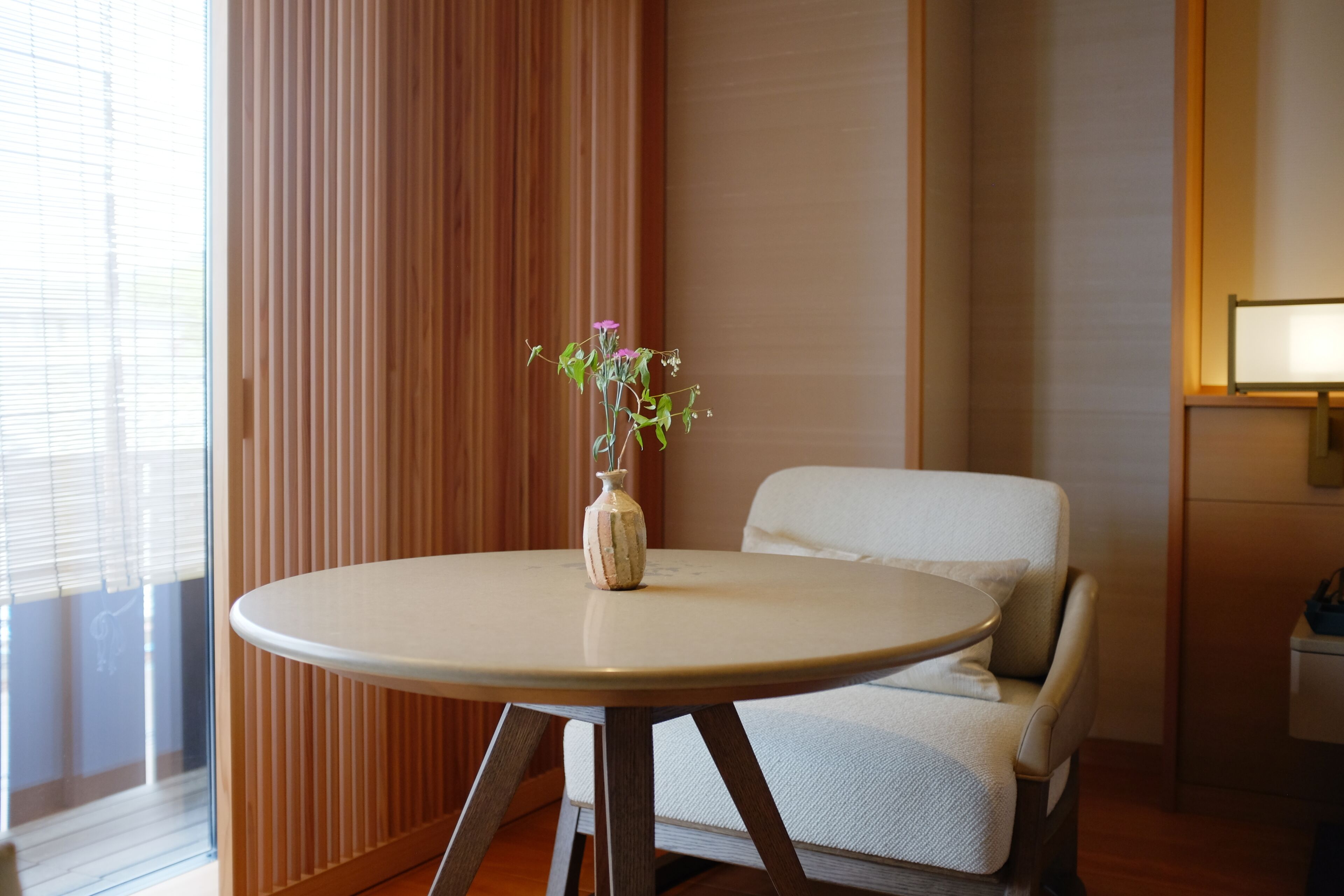 Una habitación con una mesa redonda, una silla, un florero con flores y una ventana con persianas.